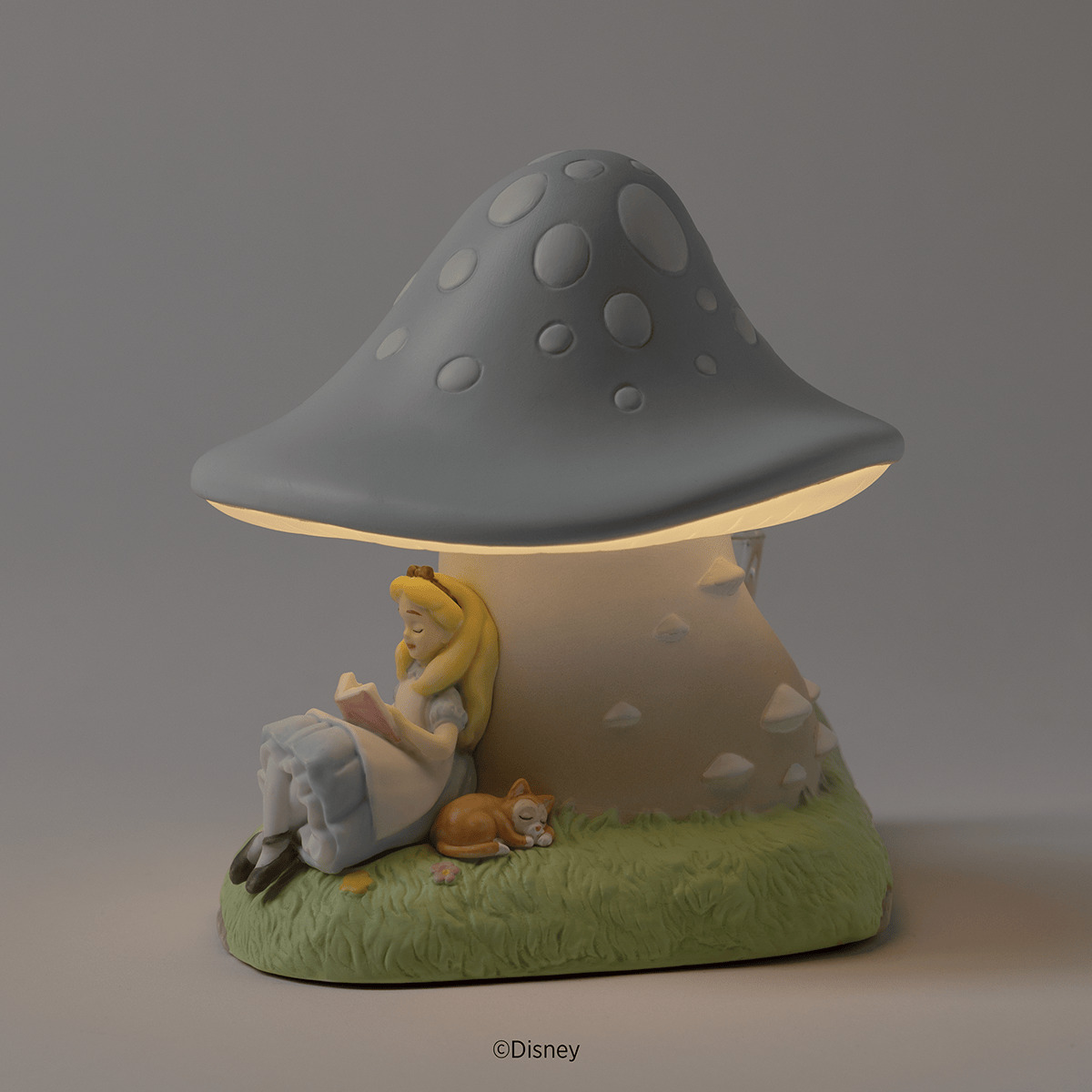 Glowing ceramic mushroom warmer with a spotted blue-grey cap, Alice asleep reading against its stem beside a curled-up orange kitten