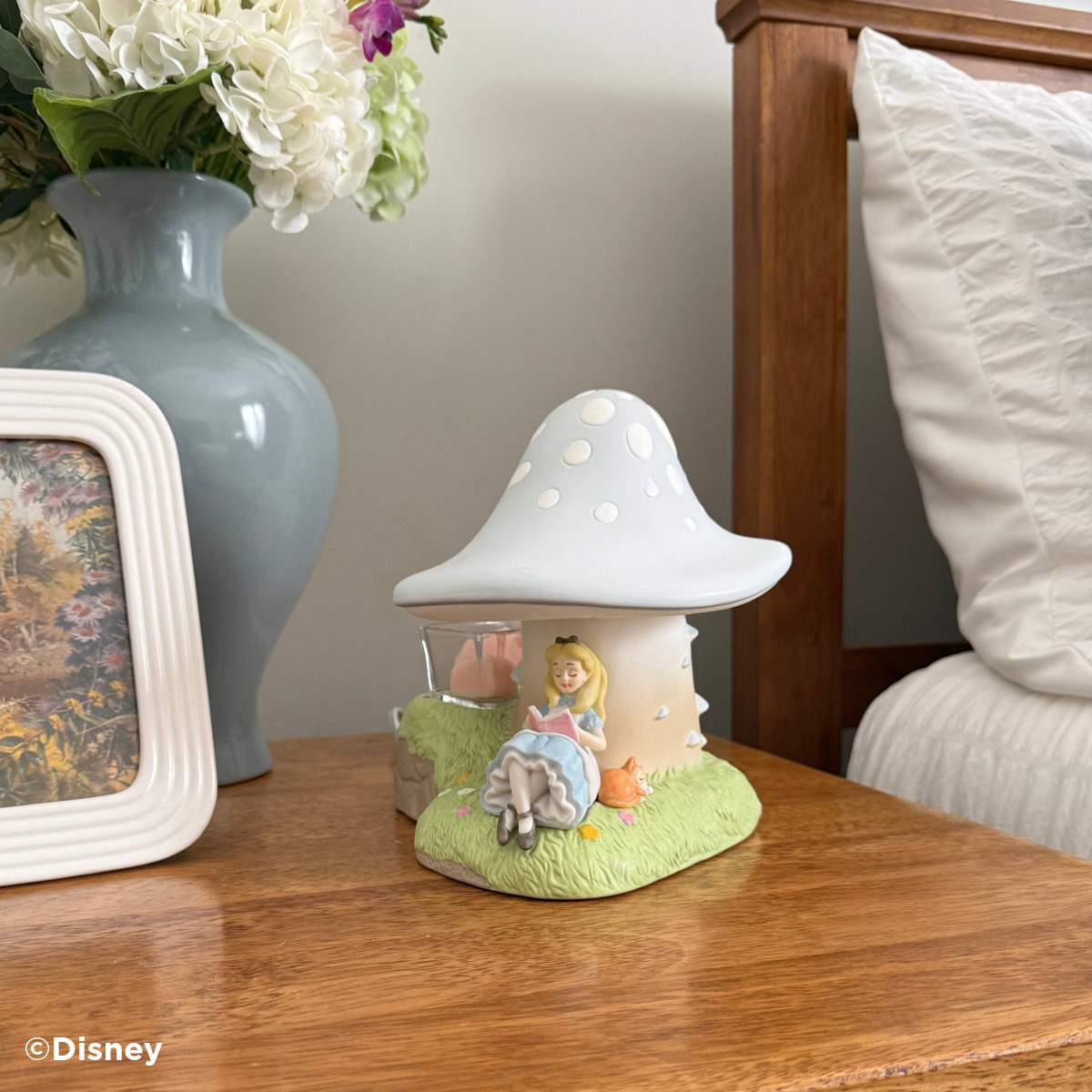 Ceramic warmer shaped like a pale blue spotted mushroom with Alice reading a book against its stem, on a bedside table
