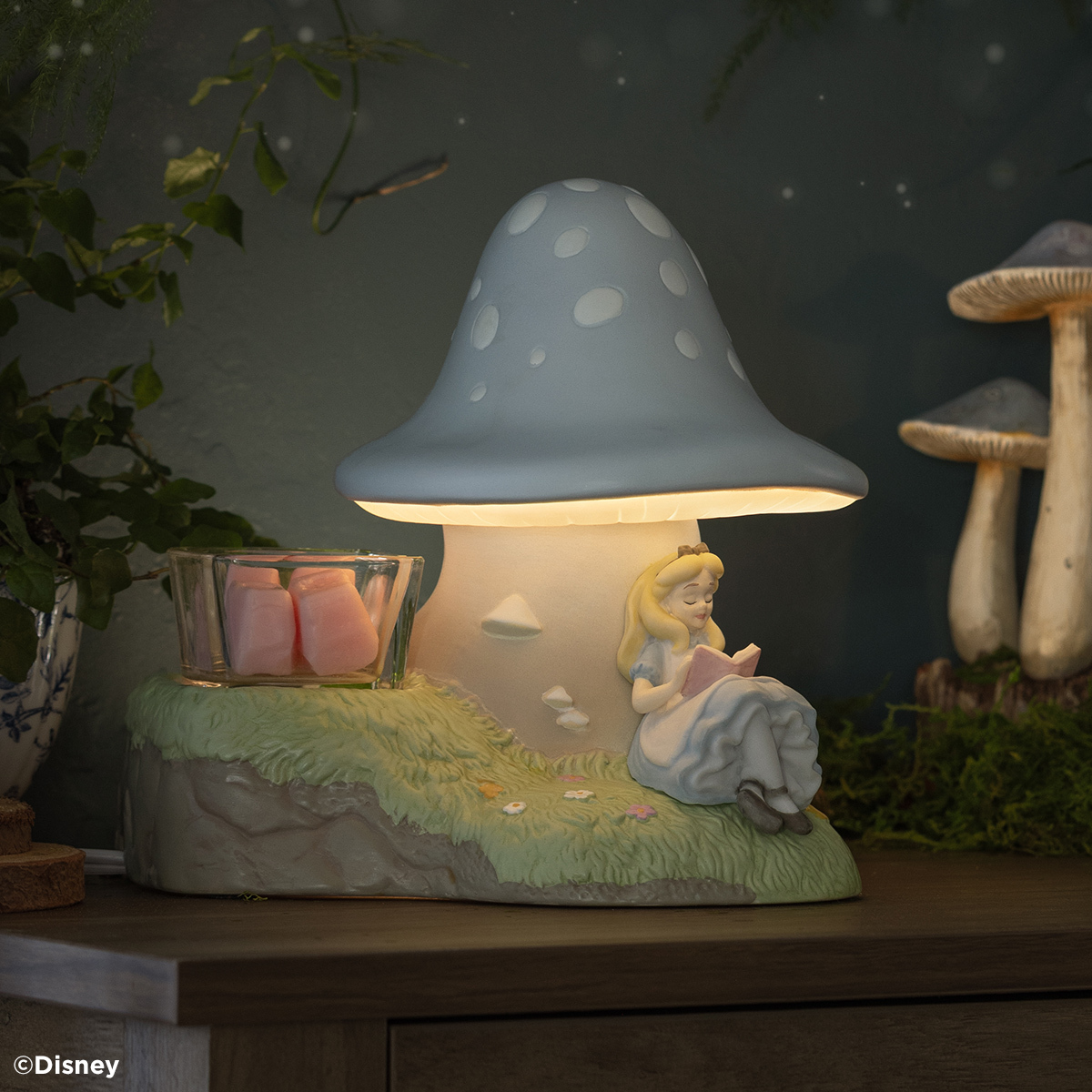 Glowing blue spotted mushroom warmer with Alice reading a book on its grassy base and pink wax cubes in a glass dish
