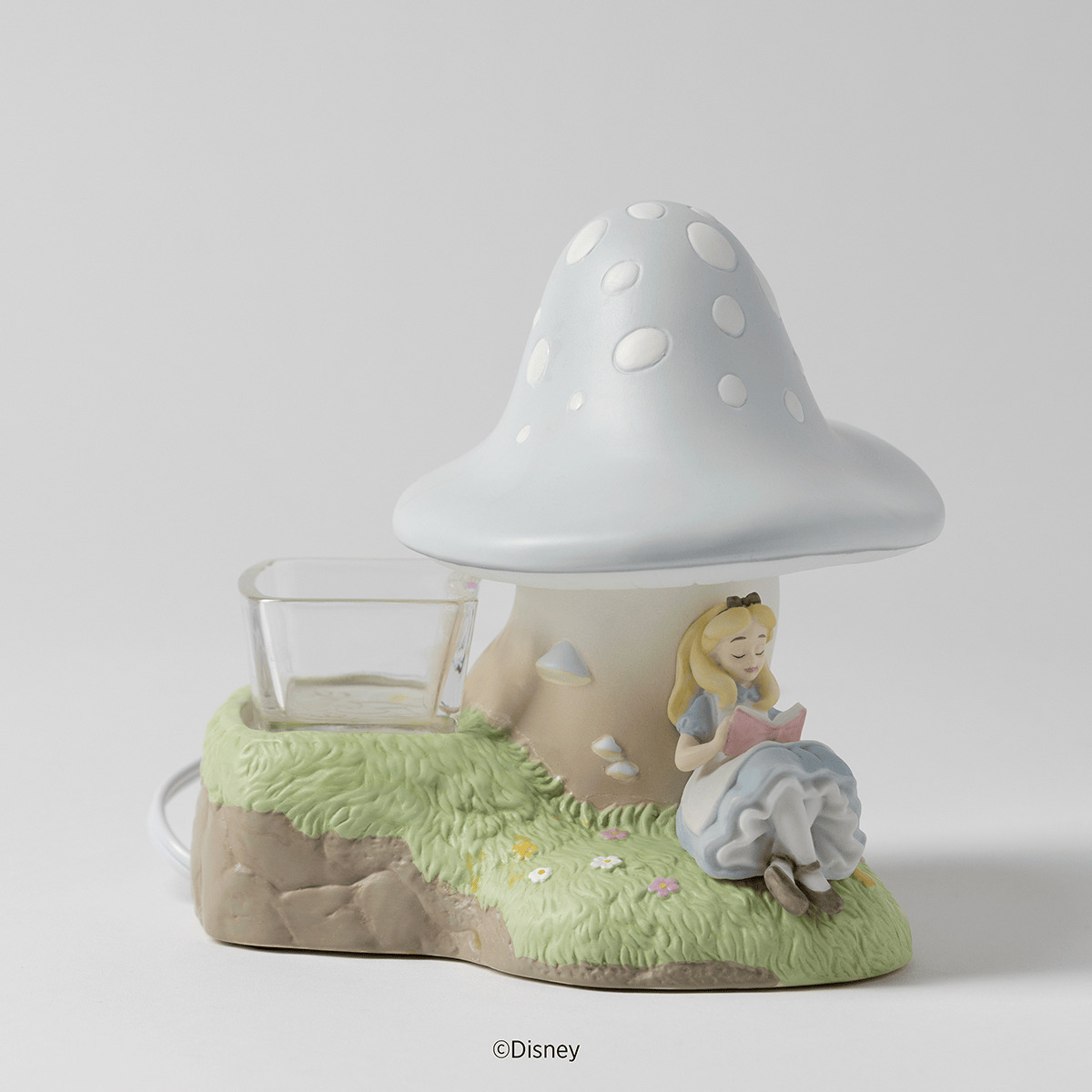 Ceramic warmer shaped as a pale blue spotted mushroom on a grassy base, with Alice reading a book beside a clear glass wax dish