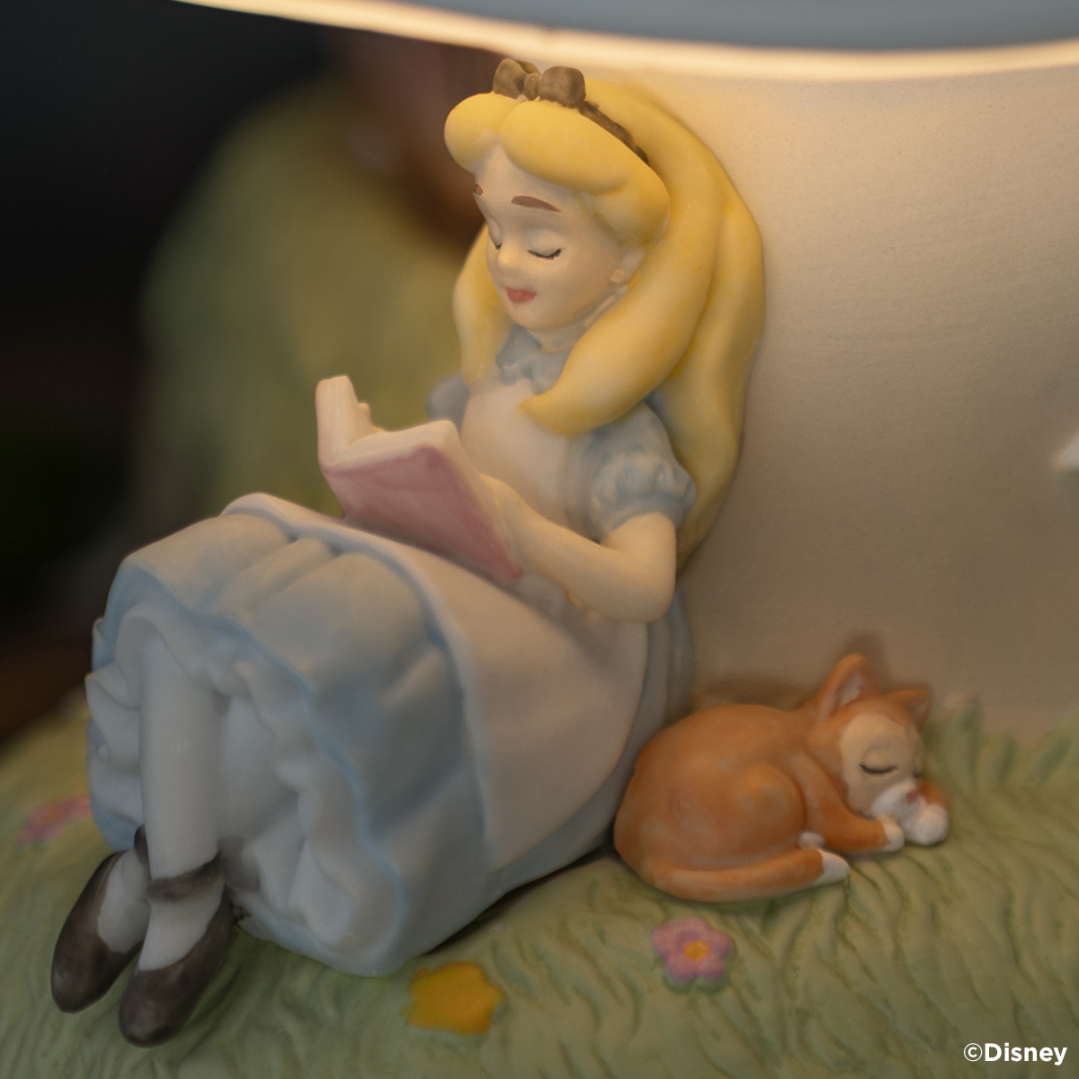 Close-up of a ceramic warmer with a sculpted Alice in a blue dress reading a pink book beside her sleeping orange kitten Dinah on grass