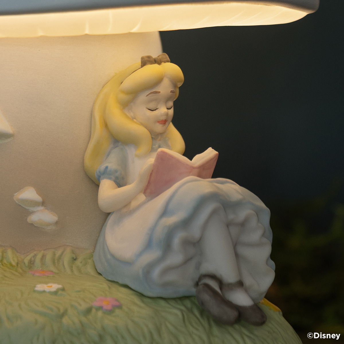 Ceramic warmer with a painted Alice figure reading a pink book, seated on grassy flowers beneath a glowing mushroom cap