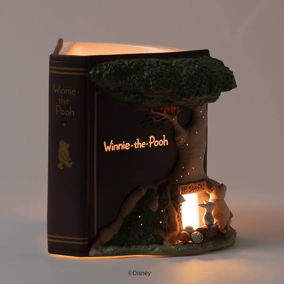 Glowing storybook-shaped warmer titled Winnie-the-Pooh, with Pooh and Piglet at a lit doorway beneath a sculpted leafy tree