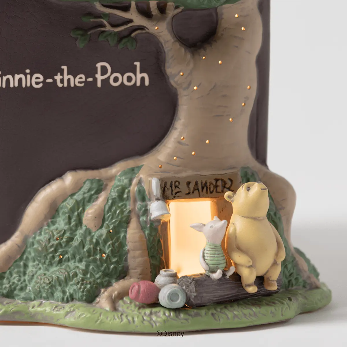 Close-up of a ceramic warmer showing Pooh and Piglet beside a glowing doorway marked Mr Sanders in a sculpted tree trunk