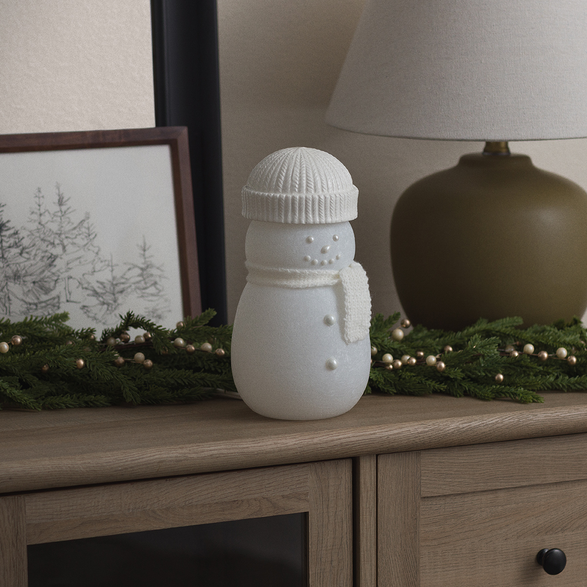White snowman warmer with a ribbed knit beanie, scarf and pearl face and buttons, on a wooden sideboard beside pine garland