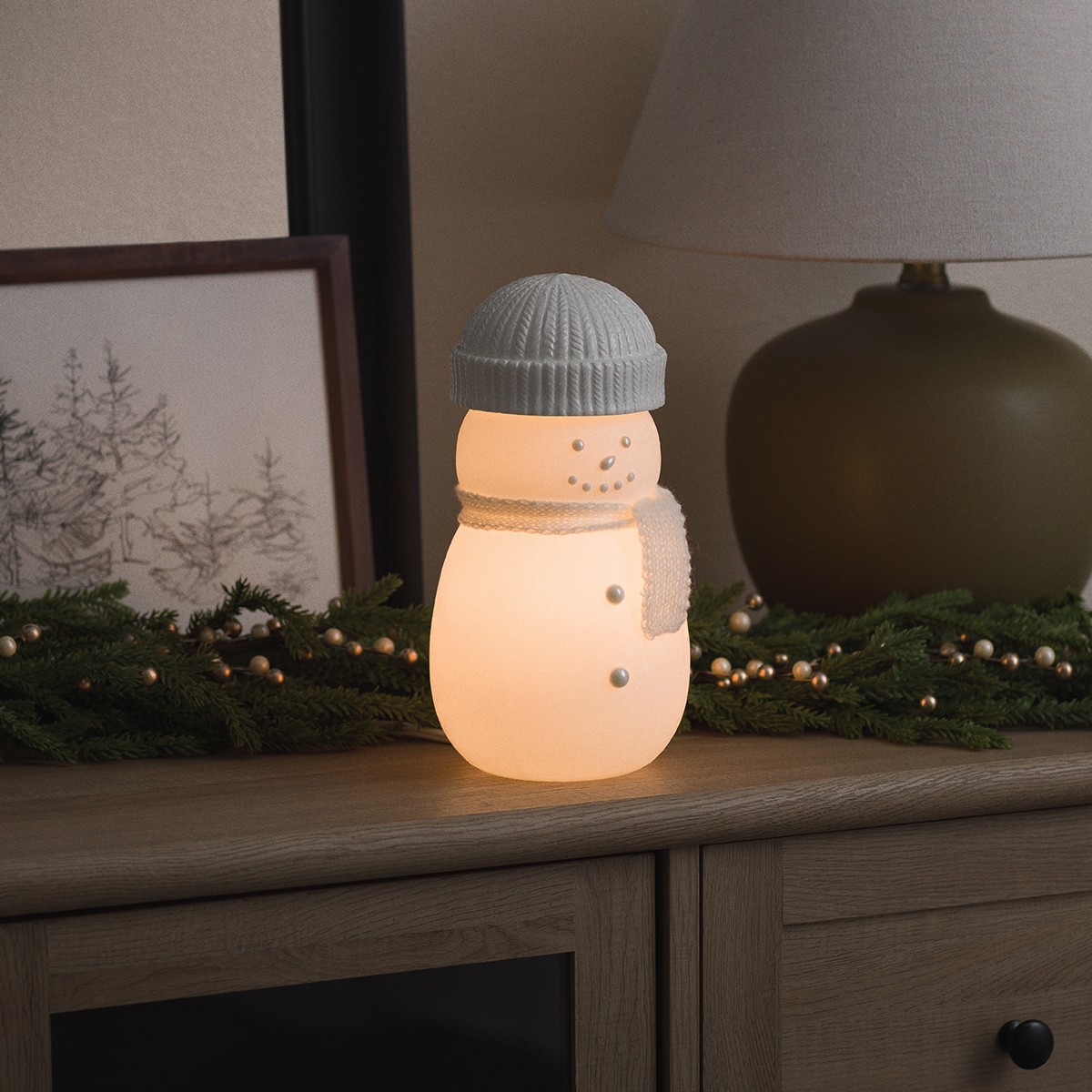 Glowing frosted glass snowman warmer with a knit beanie and scarf on a wooden sideboard beside pine garland, the Frosty Charm Scentsy Warmer