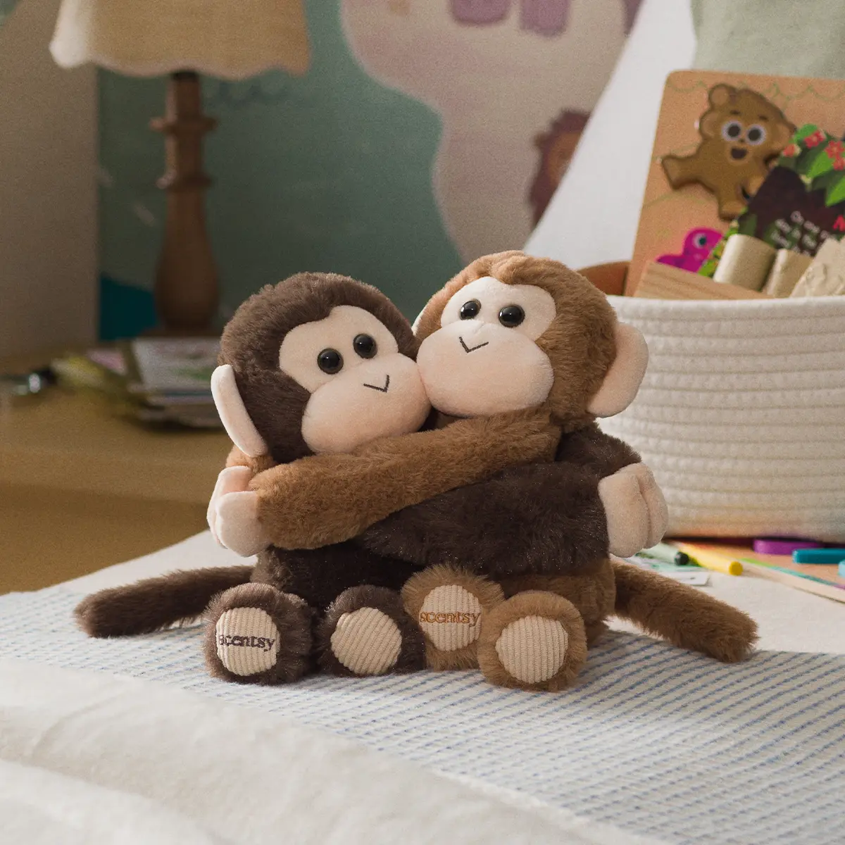 Two plush monkeys in dark and light brown hugging each other, with long tails and embroidered feet, sitting on a striped blanket