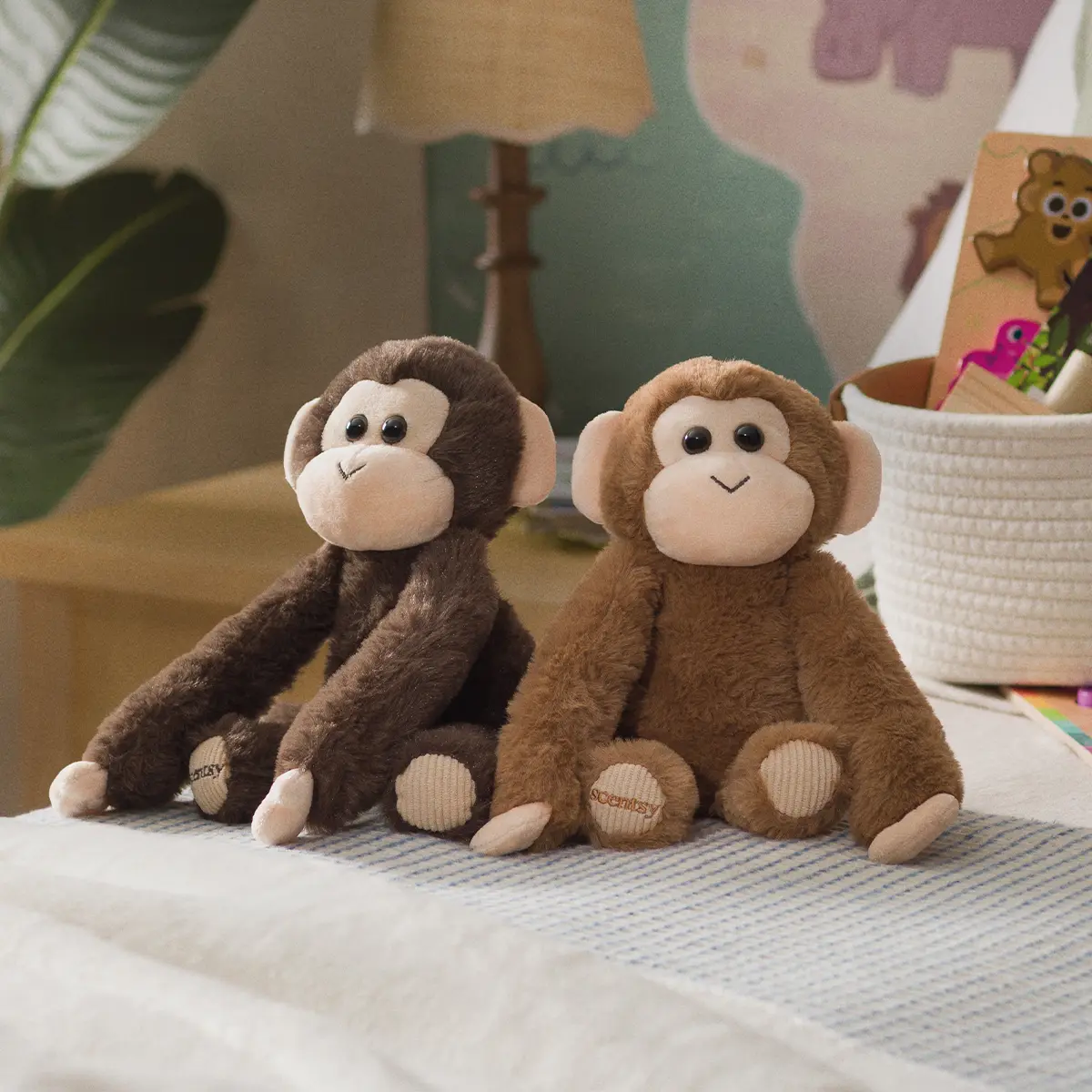 Dark brown and light brown plush monkeys with cream faces and corduroy foot pads, sitting side by side on a bed in a nursery