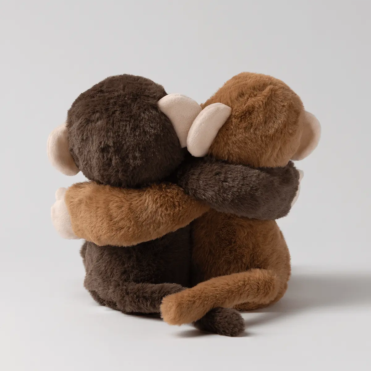 Back view of two plush monkeys, one dark brown and one tan, hugging with arms around each other and tails intertwined