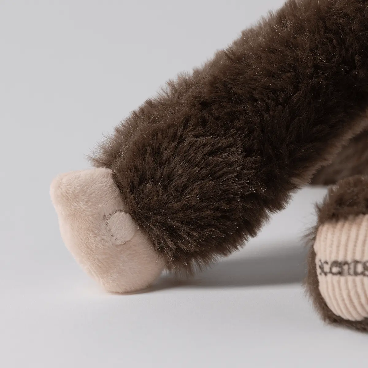 Close-up of a brown plush toy's fluffy arm with a beige velour hand, beside a ribbed corduroy foot pad embroidered with Scentsy